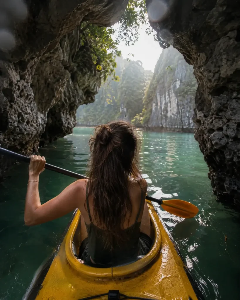 View from behind a solo traveler kayaking through a limestone arch into a hidden emerald lagoon in Lan Ha Bay near Cat Ba Island, Vietnam.