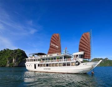 A female independent traveler standing on a classic wooden cruise boat deck looking at limestone mountains in Lan Ha Bay, Vietnam.