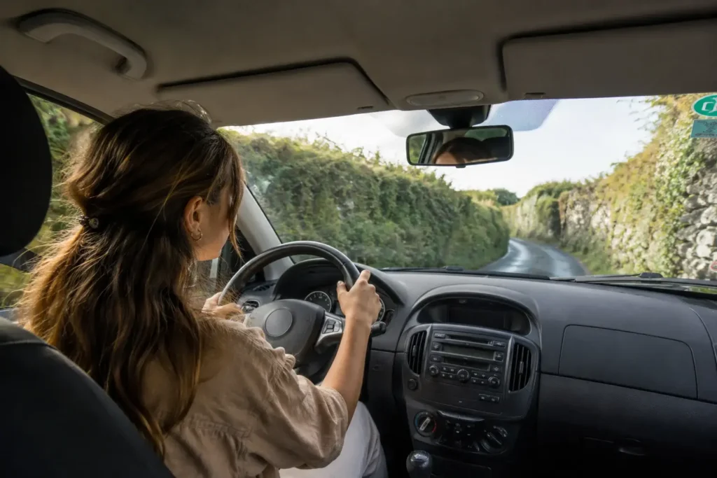 A female traveler drives a compact rental car down a narrow, winding rural regional road flanked by tall green hedges and stone walls in the Irish countryside.