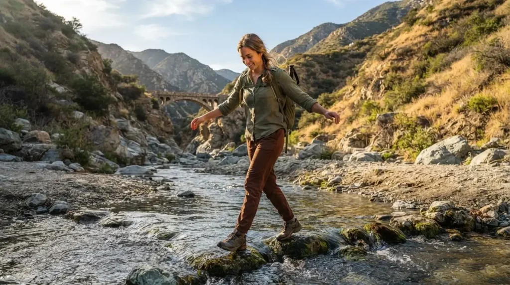 Solo female traveler crossing a rocky river ford on the trail to Bridge to Nowhere in Azusa Canyon San Gabriel Mountains California, worn boots and a small daypack, golden afternoon light